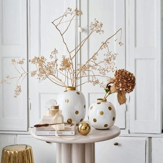 a grouping of round white vases with gold dots on a white marble side table that features other things like books, baubles etc. The vases have dried flower arrangements like hydrangeas and the room back ground is white with old fashioned edwardian shutters