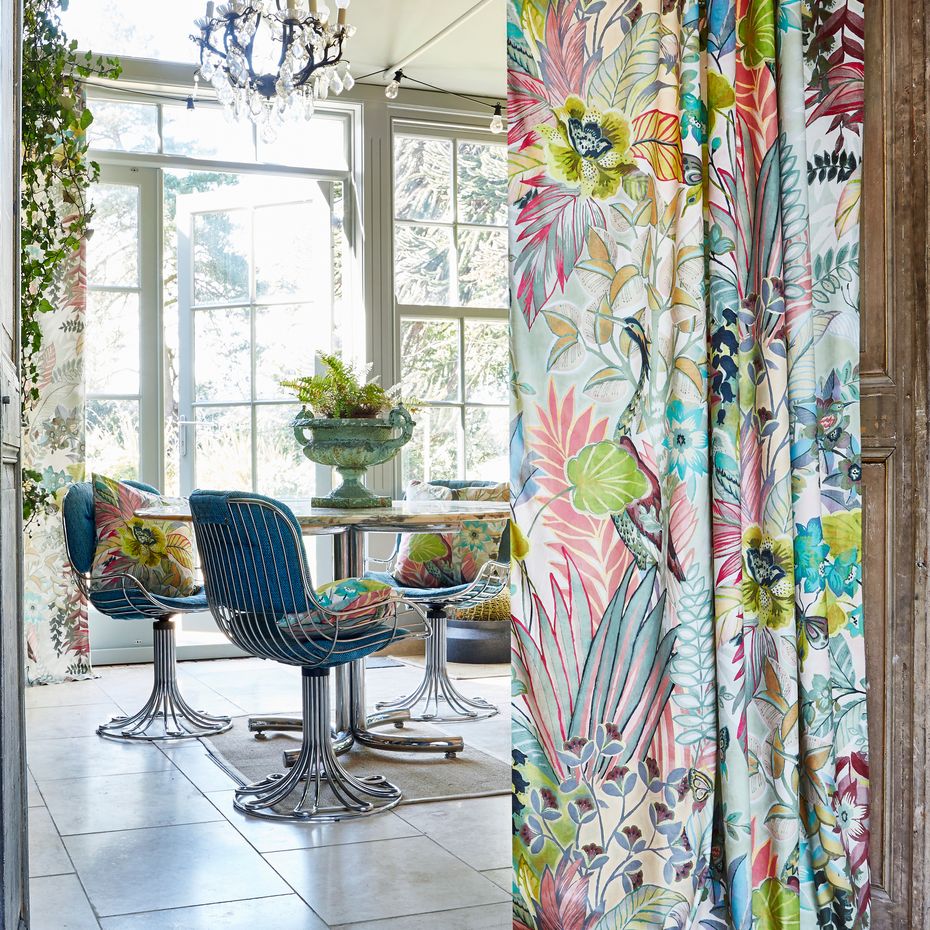 Botanist Fabric, large floral, leaves and fronds in curtains hanging in a conservatory with a dining setting with silver wire chairds and botanist fabric as cushions all sitting around a round talbe with white tiled floor