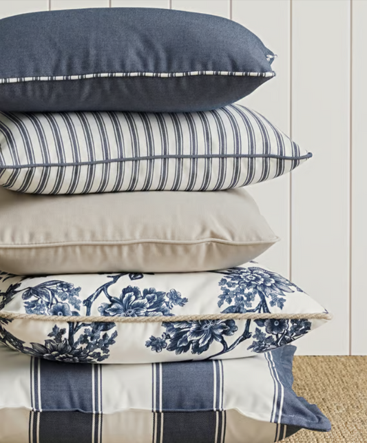 stack of cushions on a beige carpet against a white shiplap wall, indigo blue with blue and white trim, blue and white thin stripe with blue piping, stone, white with blue botanicals and white piping, blue and white large striped