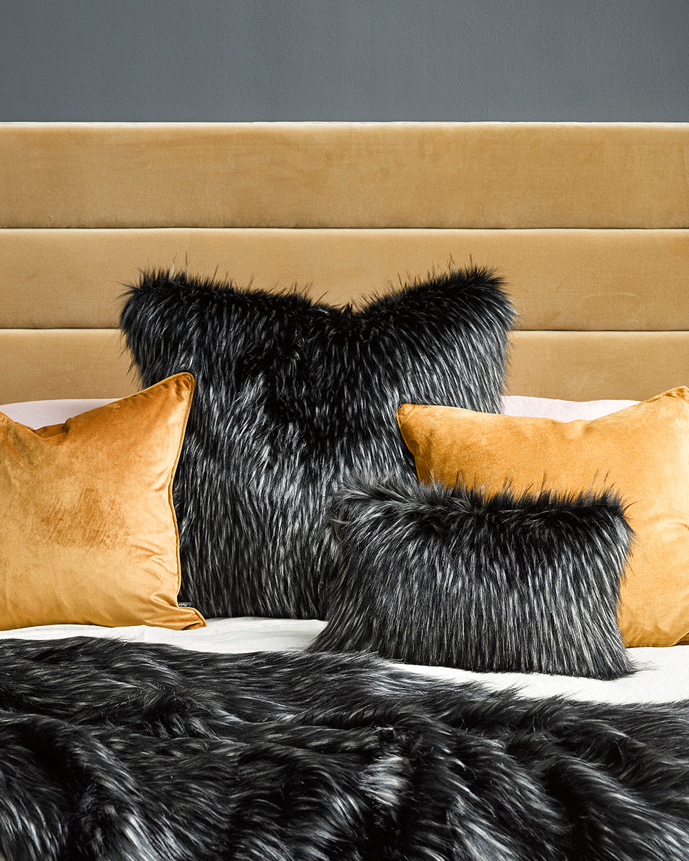 Ebony Plume imitation fur throw on bed with mustard padded headboard and cushions and a painting on grey wall and besdie table