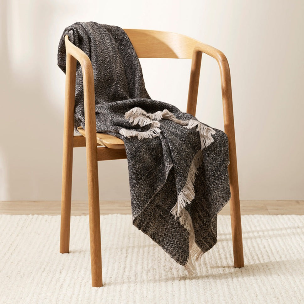 Perendale throw is a two toned black and white textured through with a white fringed edge, sitting on a plain wooden mid century style chair on a plain cream carpet