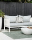 Pico outdoor cushion with deep olive tribal lines on white background placed on a grey outdoor couch with concrete plant pot with ferns, concrete round white coffee table and black cladding fence