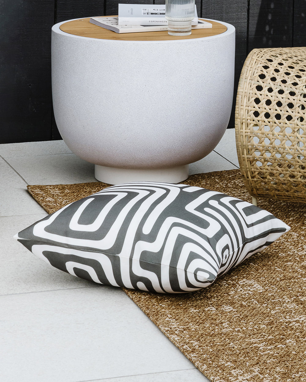 Pico outdoor cushion with deep olive tribal lines on white background placed on a brown outdoor rug with a concrete outdoor side table