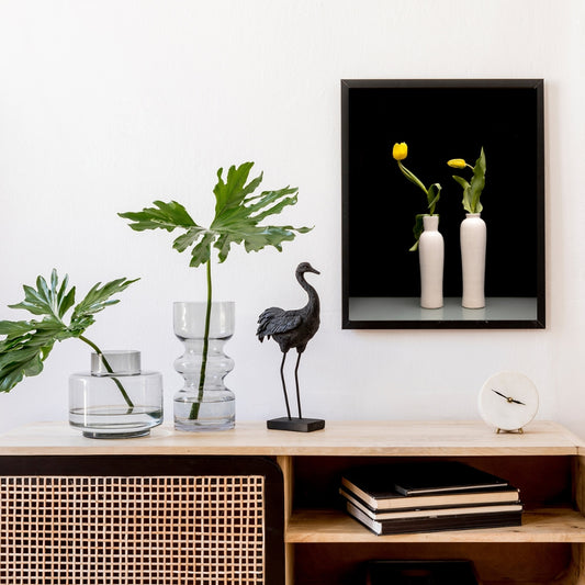 2 yellow tulips in white vases in a print sitting above a wood and rattan side board. On to pis a black moa bird statue, a white clock and two clear glass vases with green leaves