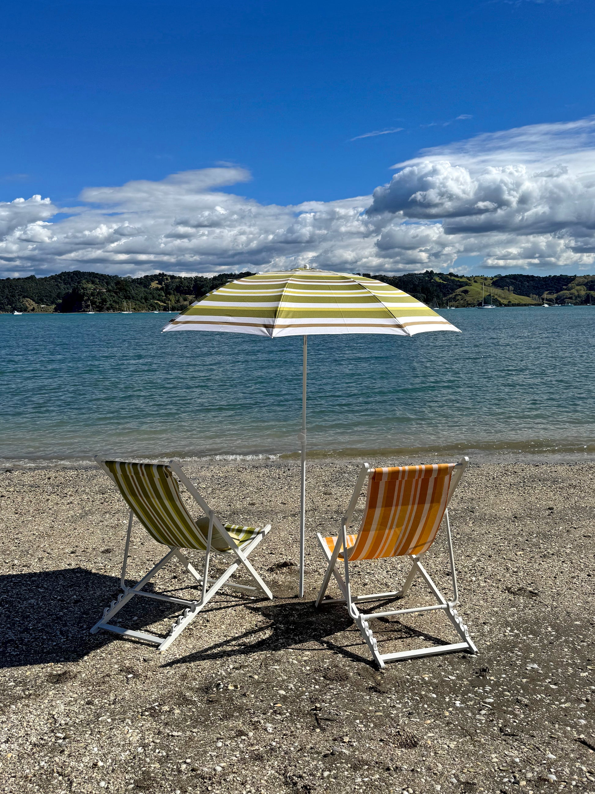 Two beach chairs under a striped umbrella on a sandy beach with ocean and sky in the background.
