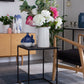 A modern black metal and glass side table in a styled living room setting, holding a white ceramic pitcher filled with white and pink flowers, alongside small blue and white ceramic vases.