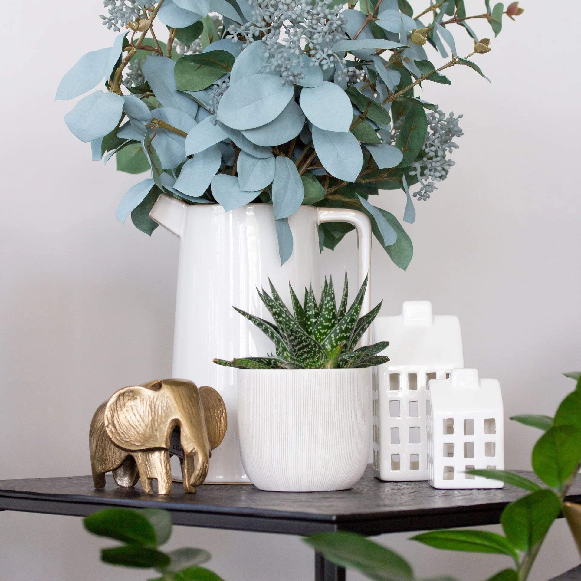 A styled tabletop arrangement featuring a large white ceramic pitcher filled with eucalyptus branches, a small white planter with a succulent, a gold elephant figurine, and white decorative house-shaped ornaments against a soft neutral background.