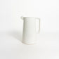 A minimalist white ceramic pitcher with a tapered body, a short spout, and a rectangular handle, photographed on a seamless white background.