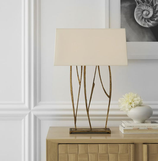 Gold lamp with tree branch design with off white parchment shade sitting on a pale wooden side board with white panelled walls behind with a amorite picture on the wall in black and white