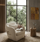 Cream arm chair with glasses and open book on it with a woodland scene of the window. Wooden and stone walls with a beige shaggy rug and wooden coffee table