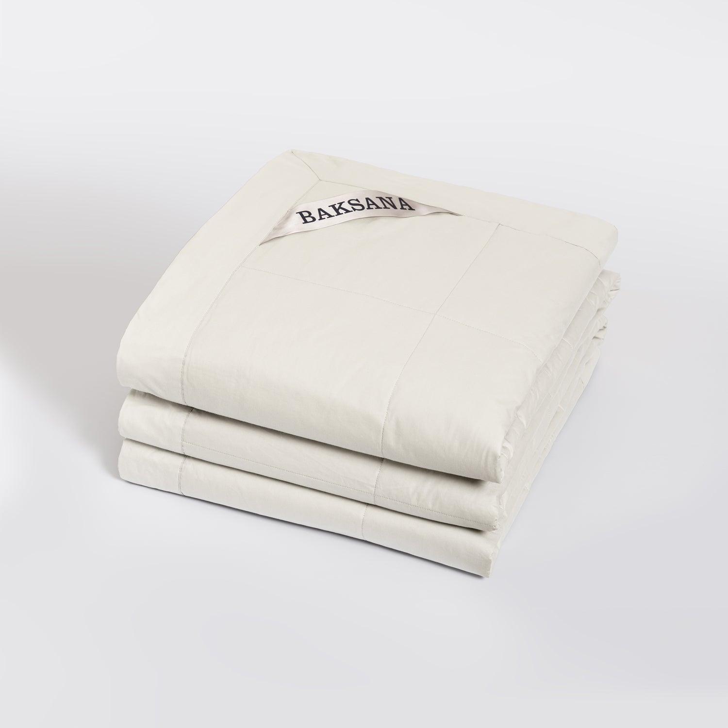A folded off-white quilted comforter or duvet with a visible BAKSANA label, photographed on a clean light gray studio background.