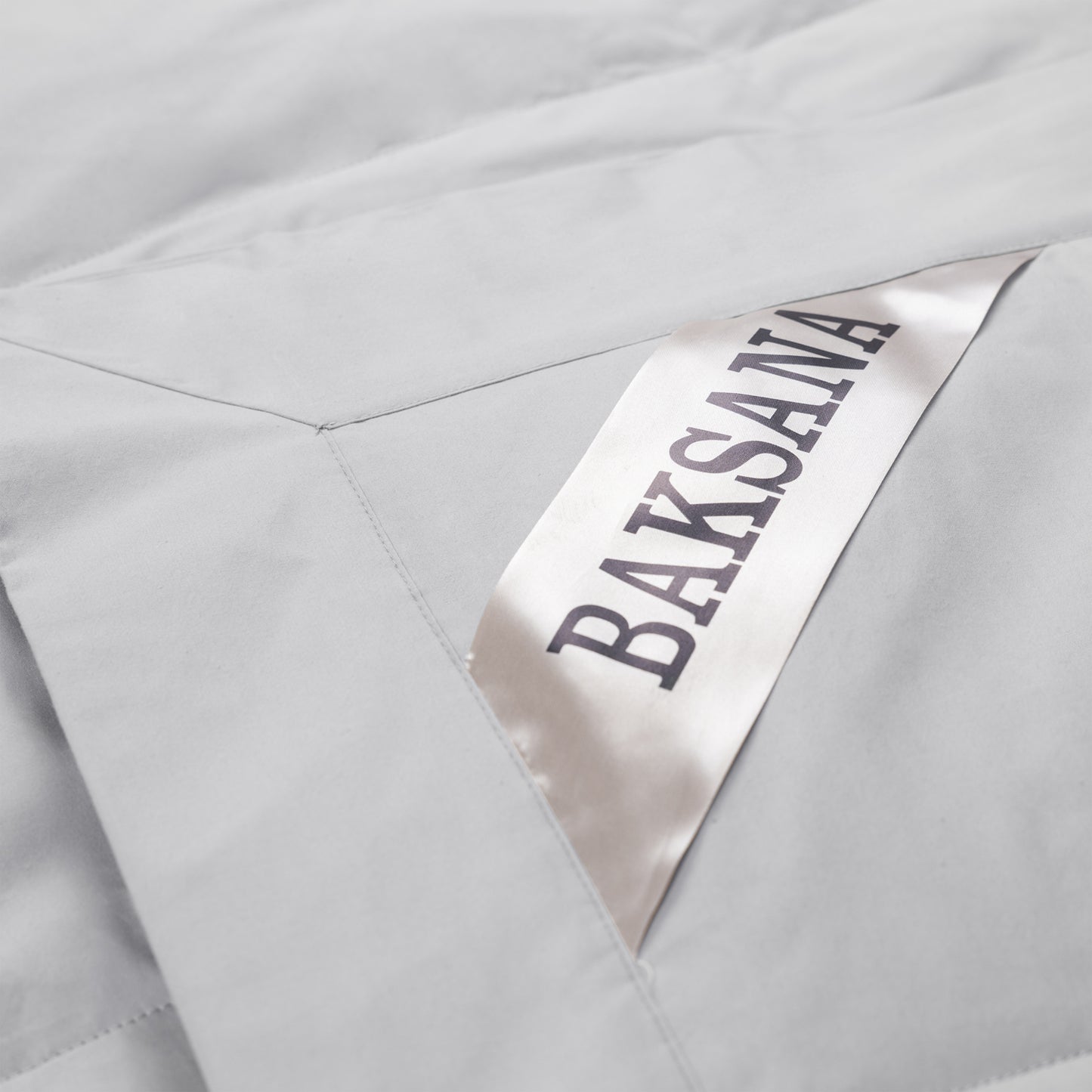 Close-up of a light gray quilted fabric with a diagonal satin label printed with the brand name 'BAKSANA' in dark lettering.
