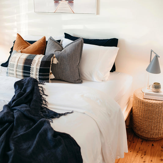 White bamboo sheets on a bed with matching throw and pillows in blues and cream and cedar with a basketweave bedside table and grey lamp shining on a coffee cup sitting on a stack of books. The walls are plain white and floor wood