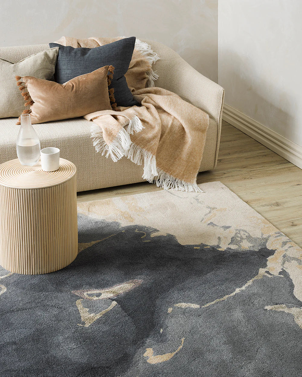 Living room with a gray and beige patterned rug, beige sofa with pillows and blanket, and wooden side table.