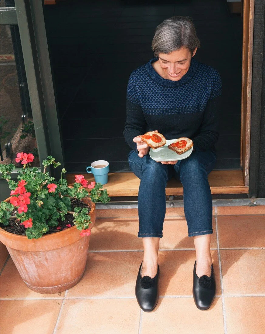 A woman sits on a wooden step in front of a doorway, holding a plate with two slices of toast topped with red jam. She is wearing a dark navy long-sleeve knit sweater with a subtle pattern, dark blue jeans, and black loafers.