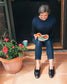 A woman sits on a wooden step in front of a doorway, holding a plate with two slices of toast topped with red jam. She is wearing a dark navy long-sleeve knit sweater with a subtle pattern, dark blue jeans, and black loafers.
