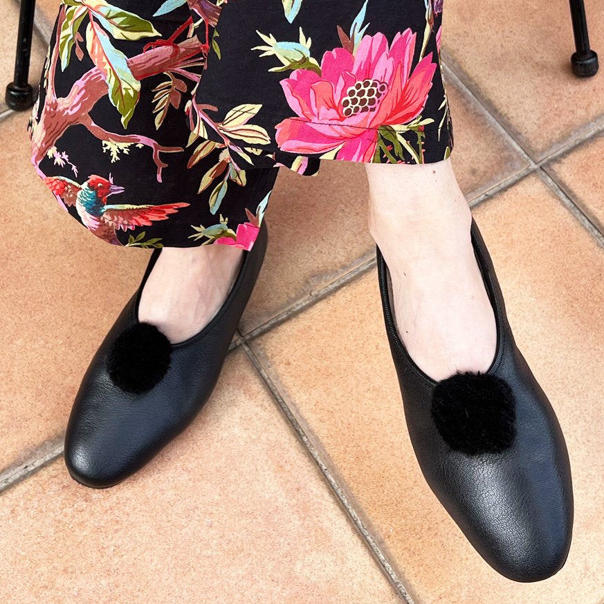 Black leather pointed-toe flats with black pom-pom embellishments, shown on a model wearing a black floral skirt on terracotta tiled flooring.