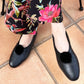 Black leather pointed-toe flats with black pom-pom embellishments, shown on a model wearing a black floral skirt on terracotta tiled flooring.