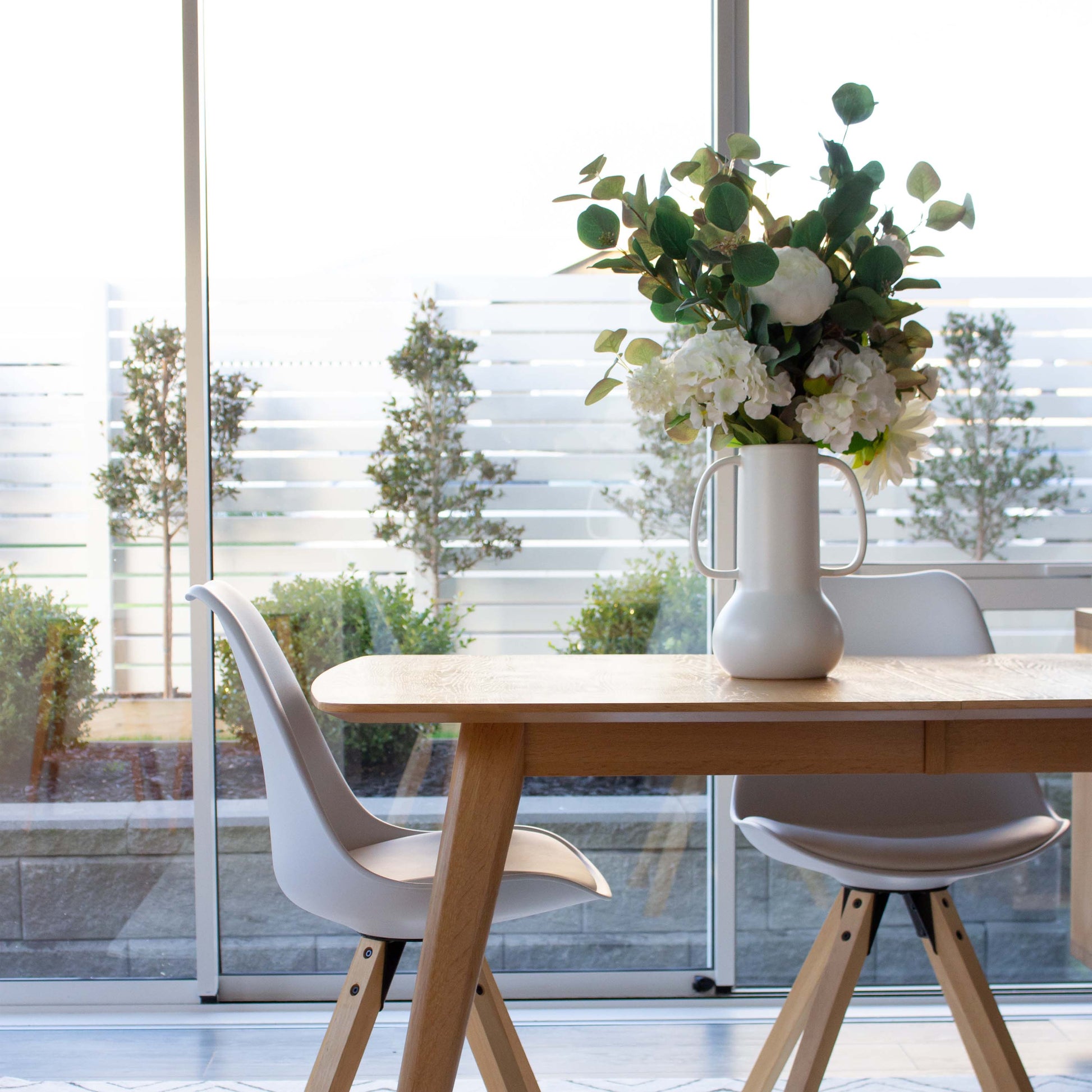 White large handled vase with white blooms sitting on a pale wooden talbe with white and wood dining chairs, window in the background