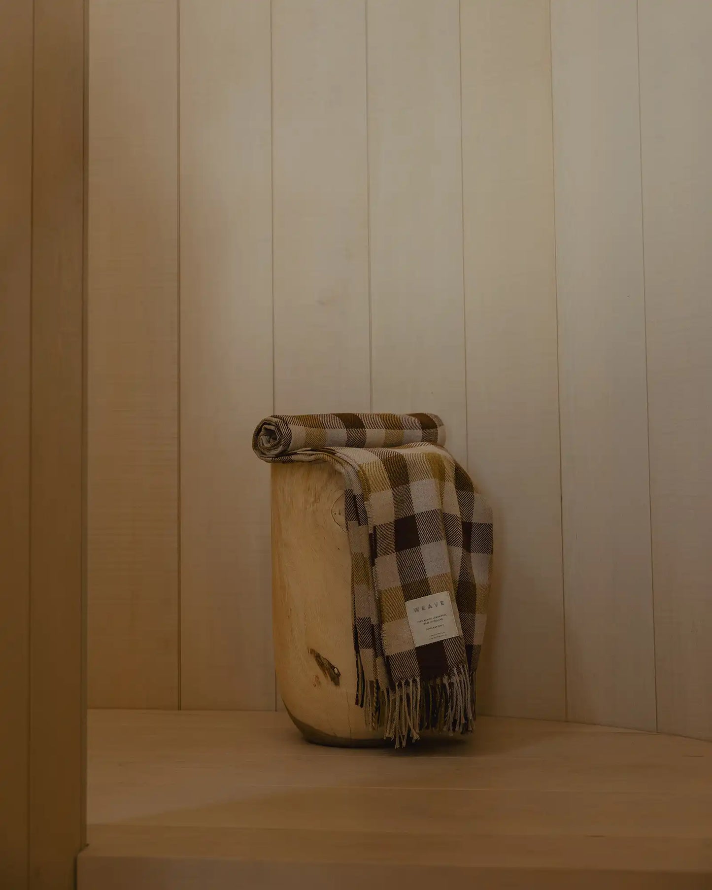 Plaid blanket draped over a wooden barrel against a wooden paneled wall.
