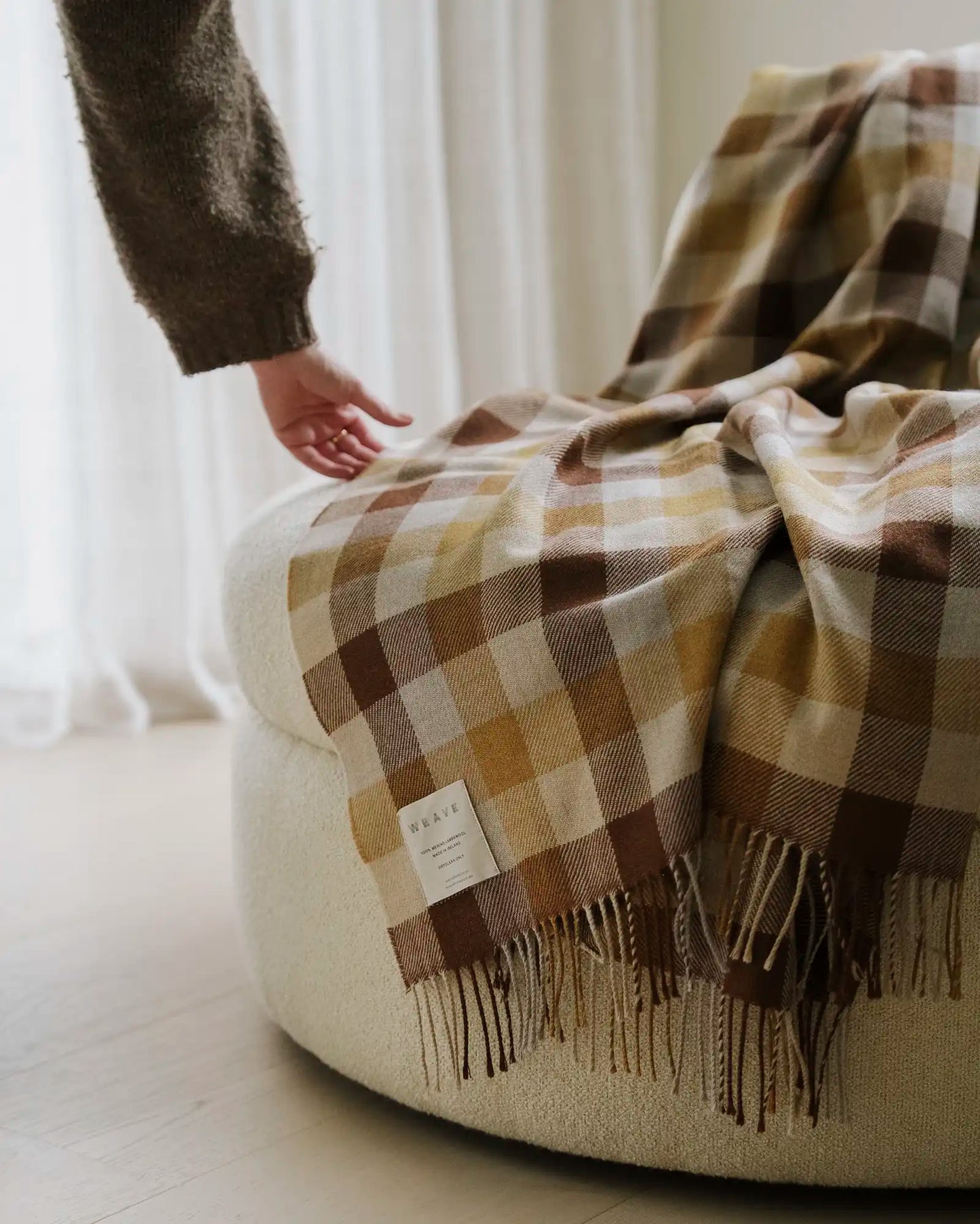 Plaid blanket draped over a chair with a blurred hand reaching towards it, set against a neutral background.