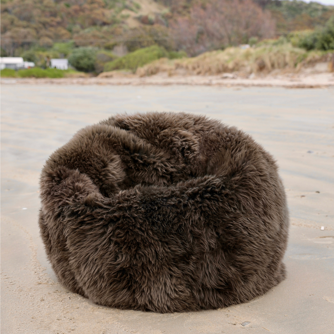 A round, plush faux-fur bean bag chair in dark brown sits on a sandy beach with dunes and greenery blurred in the background.