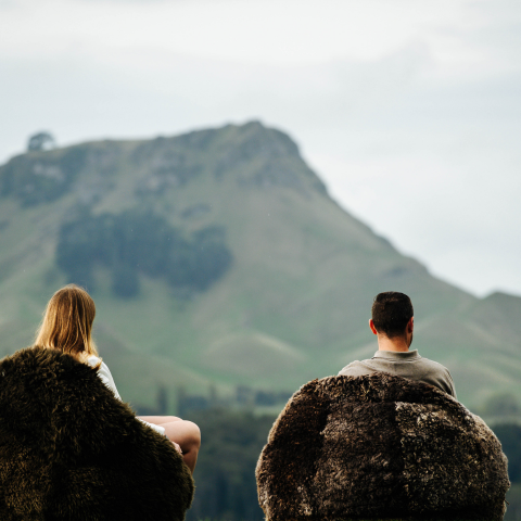 Two people sitting on large, dark, textured sheepskin beanbags, viewed from behind, looking out at a soft-focus green mountain landscape under a cloudy sky.