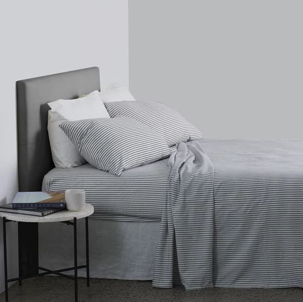 A neatly made bed with a striped duvet cover in a neutral color, accompanied by matching pillows and a small table with a book and a cup beside the bed.