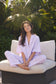Woman in lavender pajamas sitting on a white chair outdoors with greenery in the background