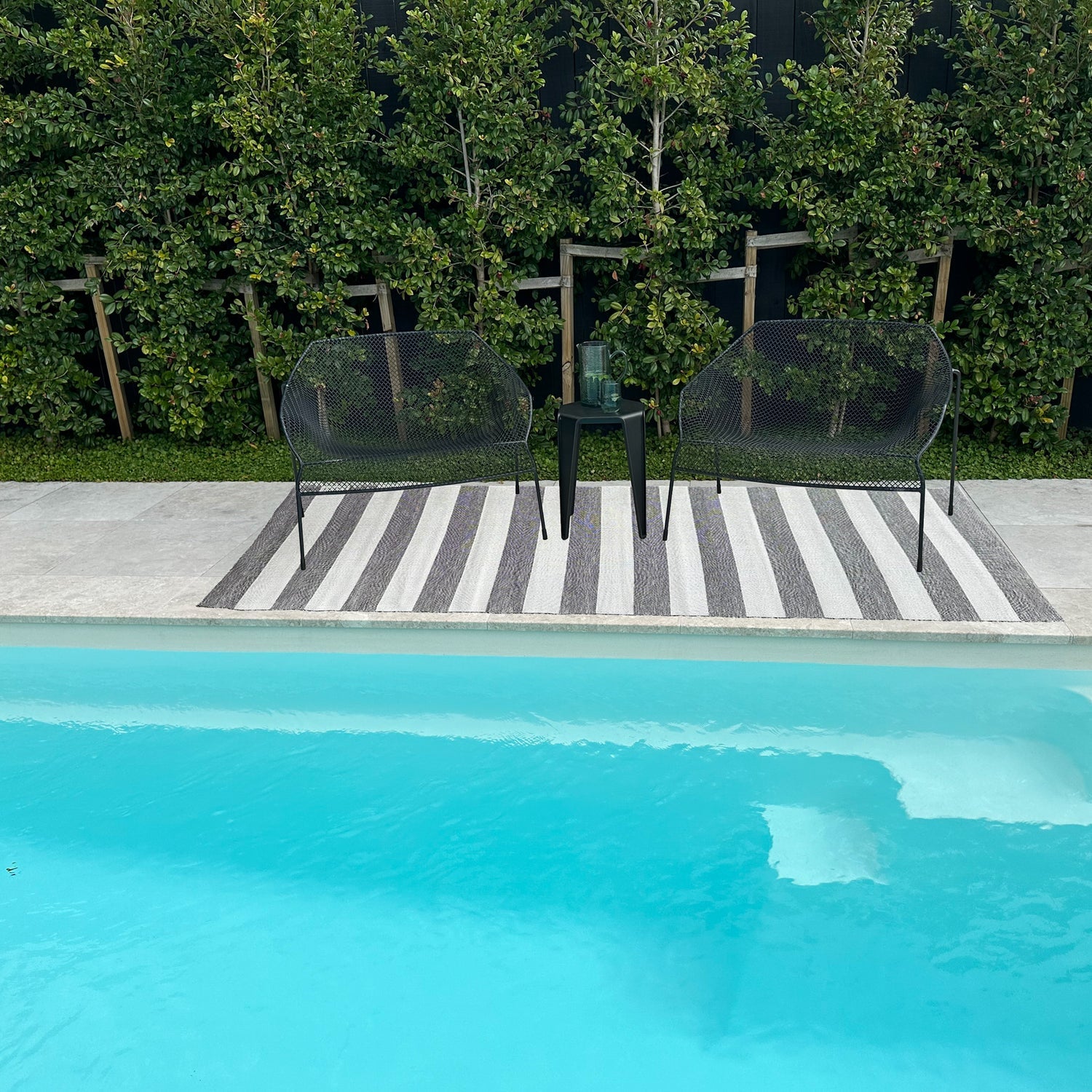 Two black chairs and a small table by a poolside with greenery in the background