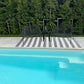 Two black chairs and a small table by a poolside with greenery in the background