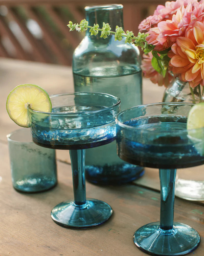 Two blue cocktail glasses with a lime wedge on a wooden table, accompanied by a bottle and flowers.
