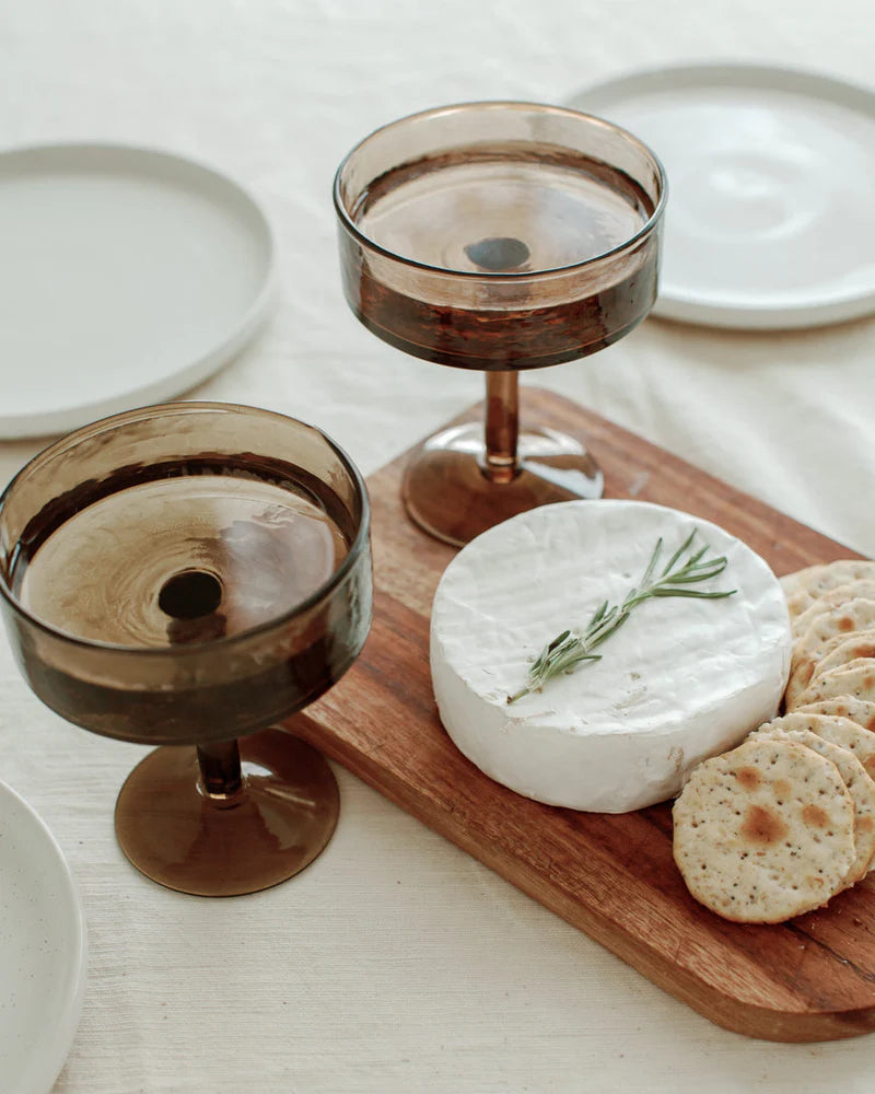 Wooden cutting board with cheese, crackers, and glasses on a white surface