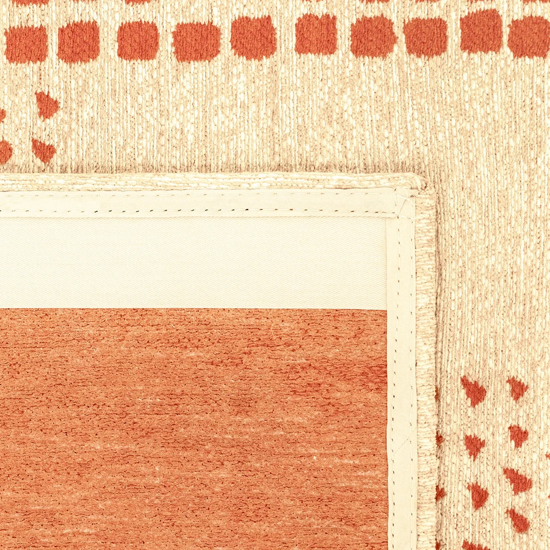 Close-up of a woven rug corner showing a cream border with orange-red geometric patterns and a matching orange-red central panel on a beige background.