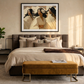 A framed art print depicting three women in flowing cream garments hangs above a bed in a styled bedroom. The print has a black frame and white mat, set against a warm beige wall.