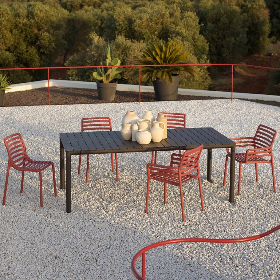 Outdoor setting with a black table and red chairs on a gravel surface, surrounded by greenery.