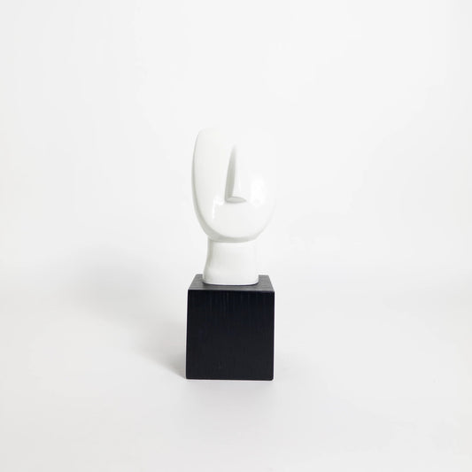 A minimalist abstract white sculpture on a black square pedestal, photographed against a seamless white studio background.