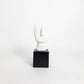 A minimalist abstract white sculpture on a black square pedestal, photographed against a seamless white studio background.