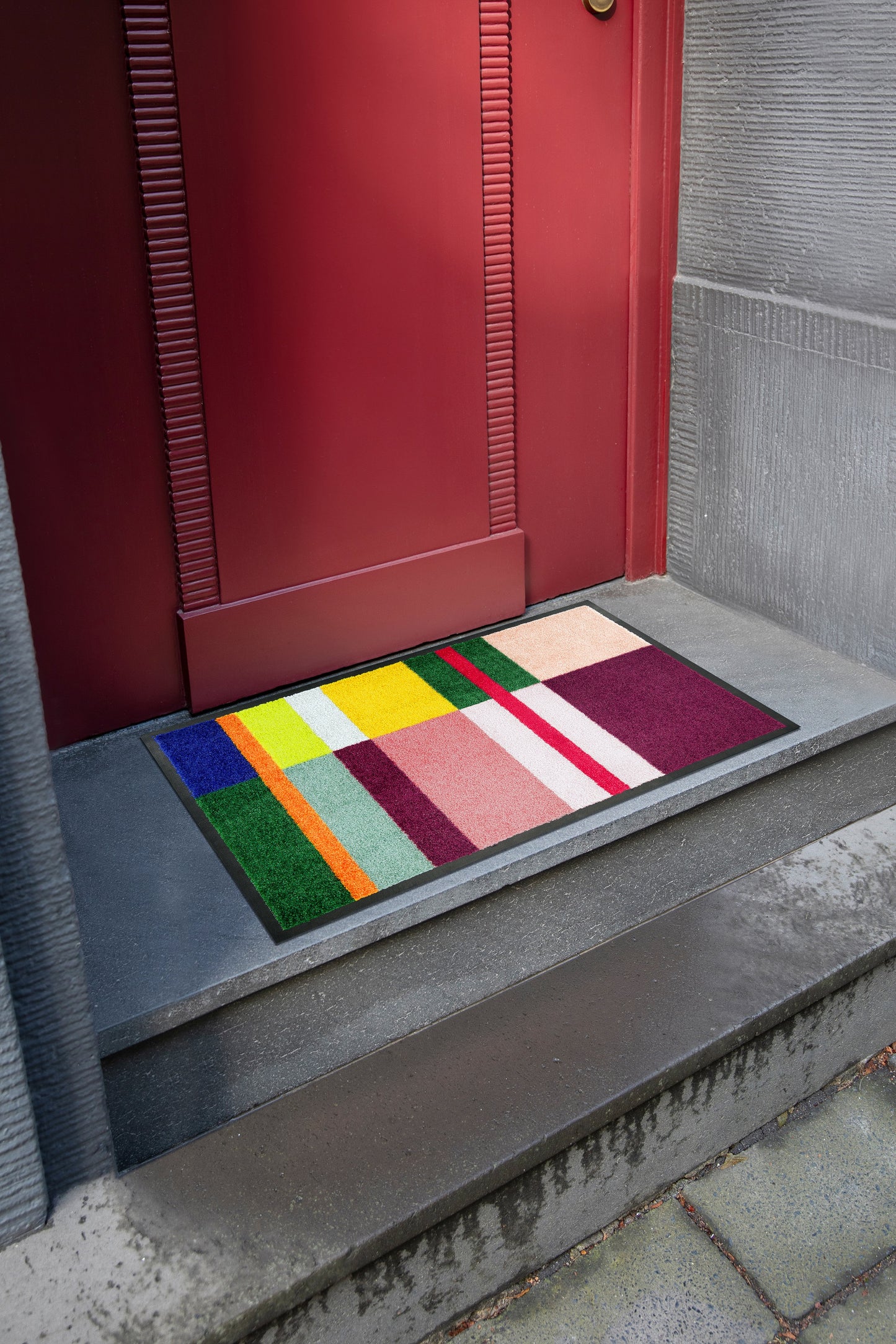 Colorful doormat with geometric pattern on a concrete step leading to a red door.