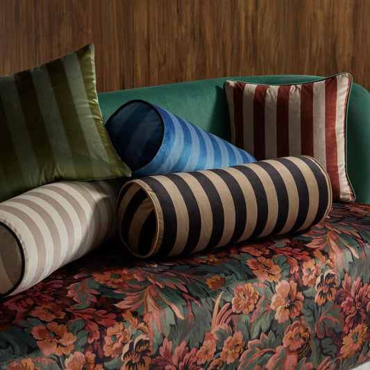 Stack of 2 square and 3 round oblong cushions in velvet stripes in blue, green, cream, black and terracotta with a plain back ground siting on a velvet floral couch with a co ordinating green back