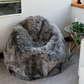 A large, plush faux-fur bean bag chair in a mottled gray and brown colorway, styled in a home interior next to a glass side table with a plant.