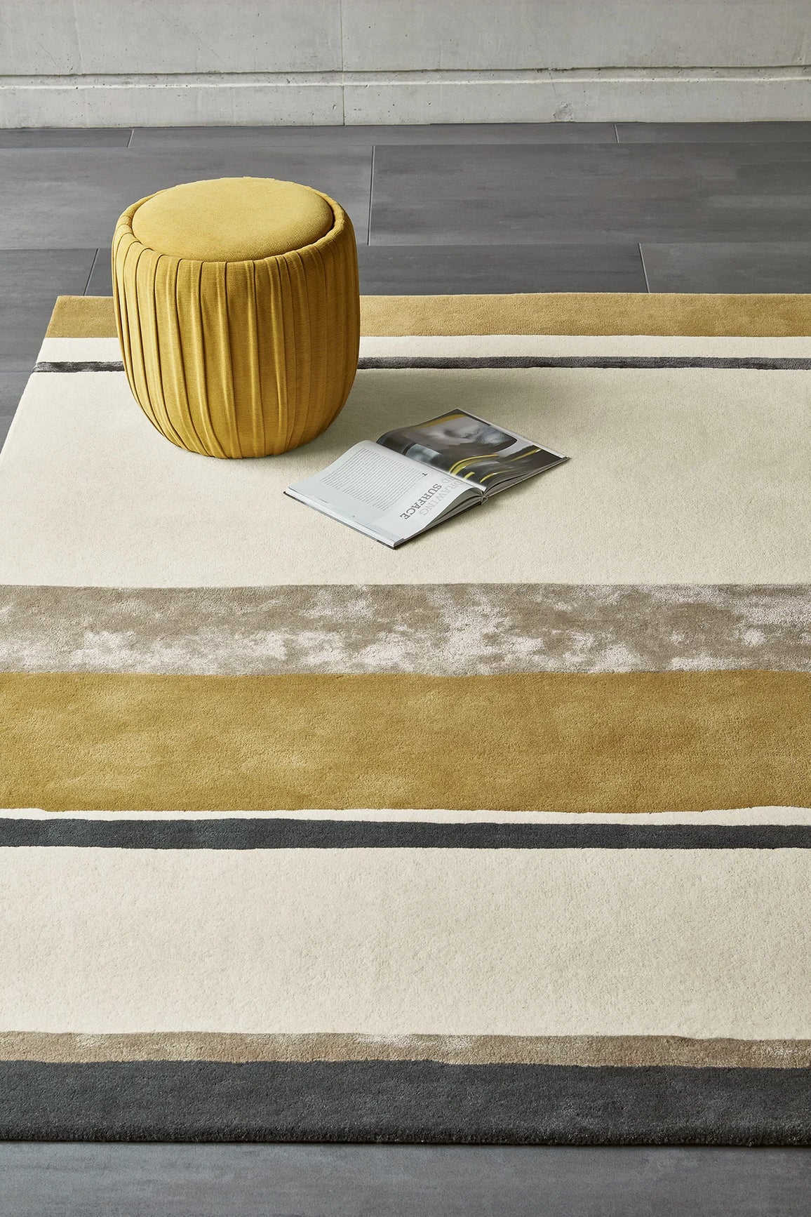 Striped rug with a yellow stool and magazine on a gray floor.