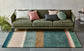 Green sofa with colorful cushions on a striped rug in a room with a plant and side table.