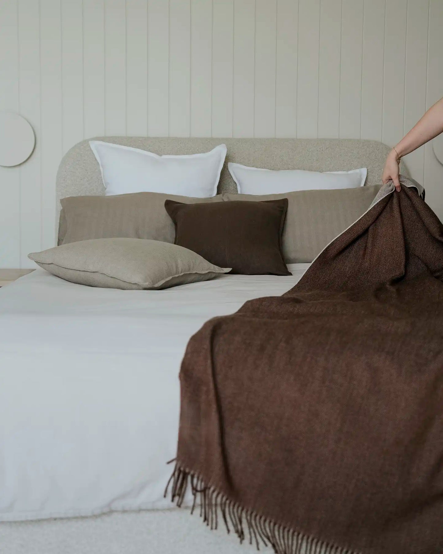 Person adjusting a brown blanket on a bed with beige and brown pillows.