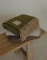 Folded green blanket with fringes on a wooden bench against a neutral background