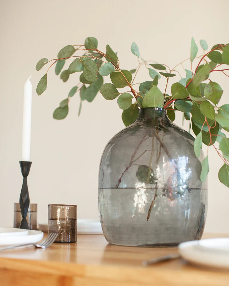 Decorative vase with greenery on a table with candles and plates.
