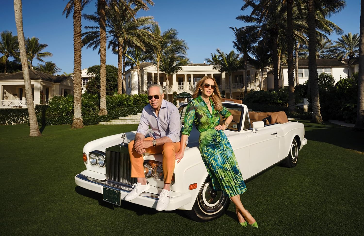 Two people sitting on a white convertible car with palm trees and houses in the background