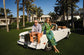 Two people sitting on a white convertible car with palm trees and houses in the background