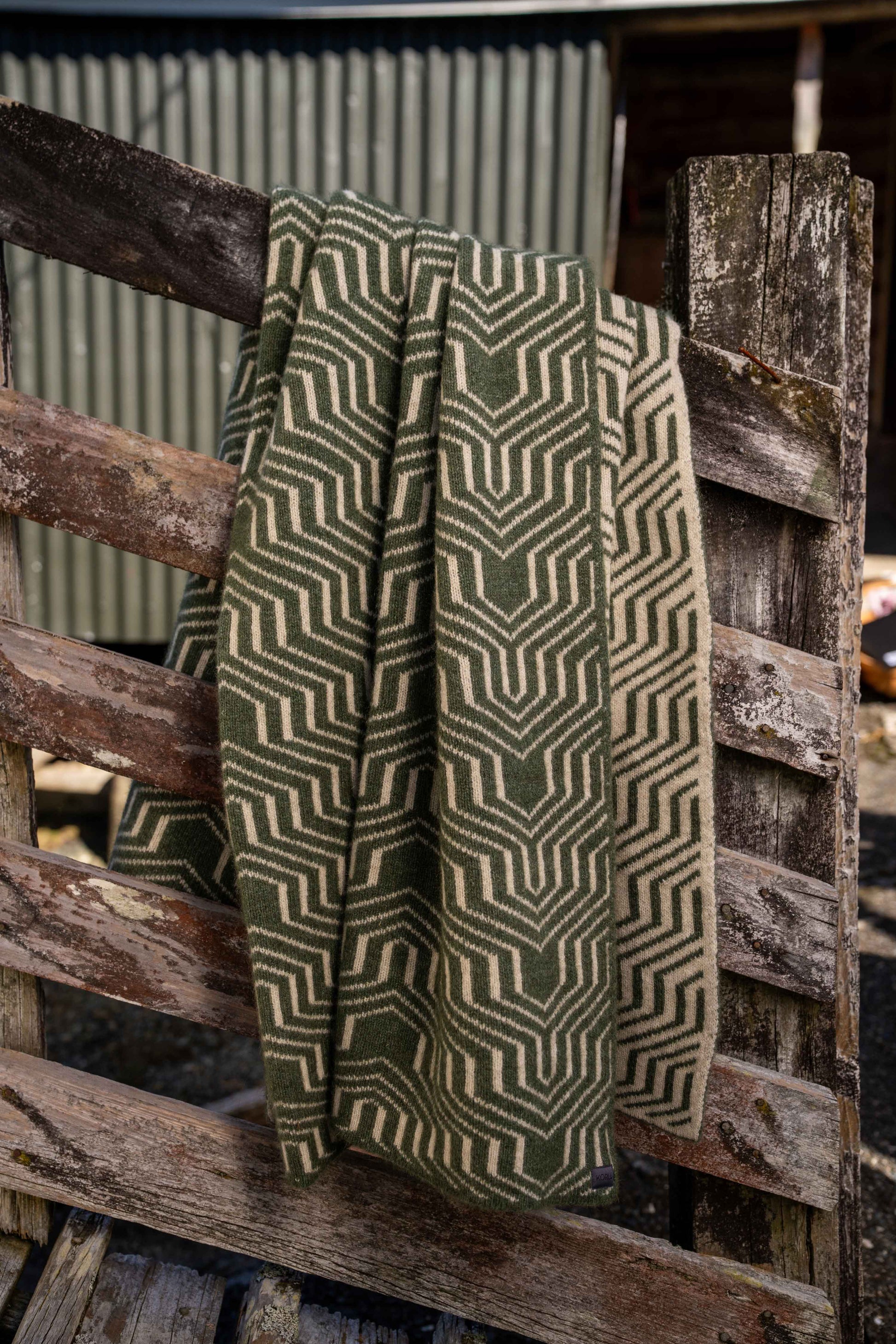 A woven throw blanket with a geometric chevron pattern in olive green and beige, draped over a rustic wooden fence.