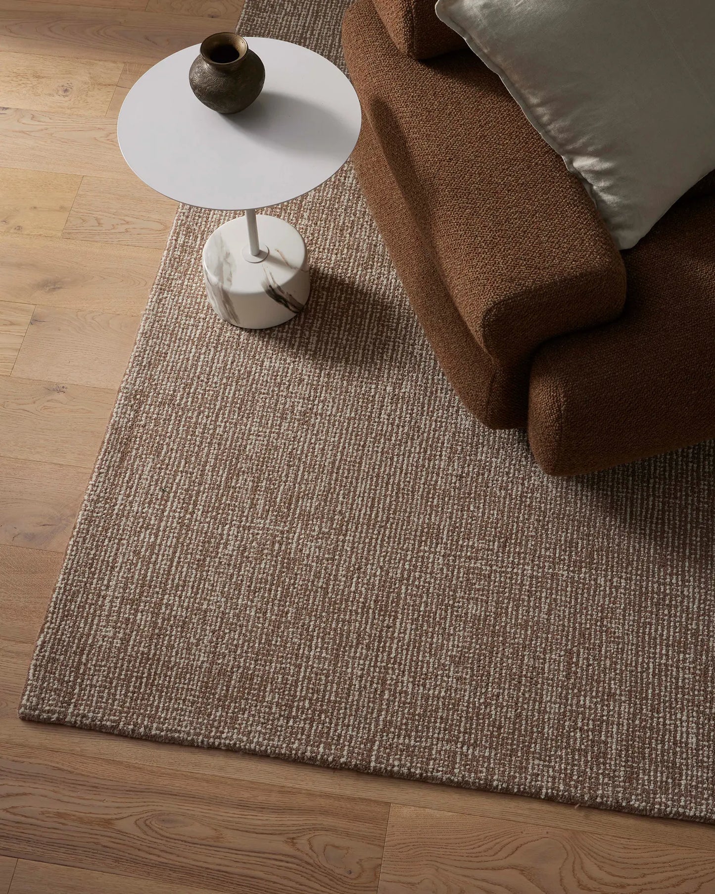 Brown textured rug on a wooden floor with a brown sofa and white side table.